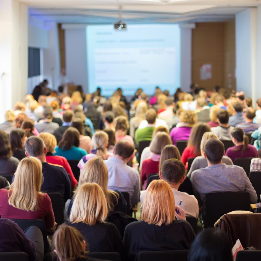 Audience seated in a conference room facing a presentation screen during a sales keynote session.